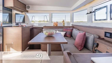 accogliente salone interno di un catamarano Lagoon 40. Il design elegante include tonalità di legno marrone, comodi cuscini rossi e grigi, e un'ampia vista sul mare attraverso le finestre panoramiche. Un ambiente perfetto per rilassarsi dopo una giornata al sole.
